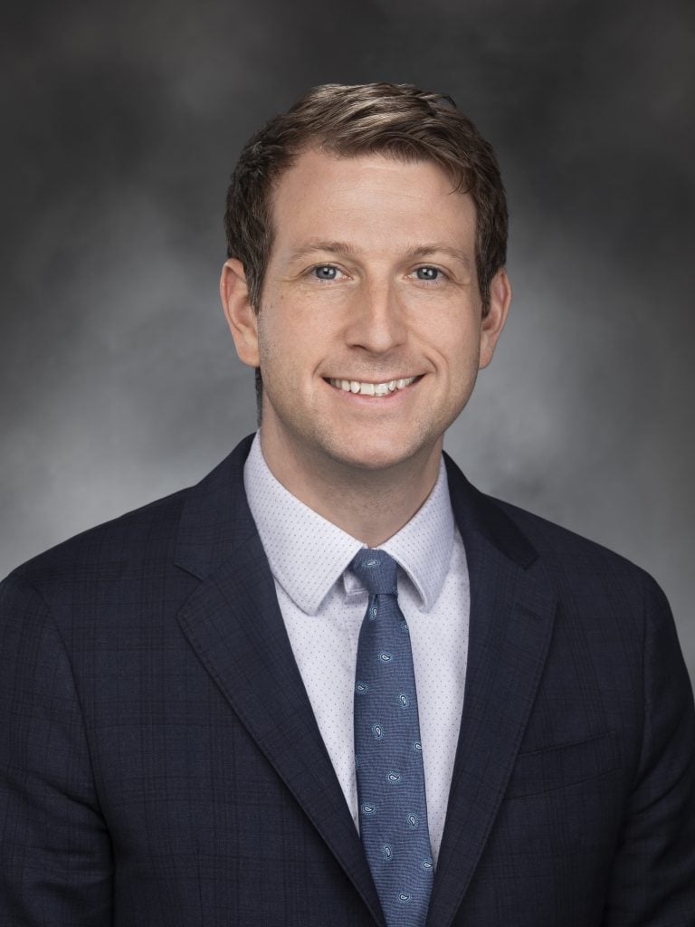 A headshot of Joe Fitzgibbon smiling at the camera while wearing a suit in front of a gray backdrop.